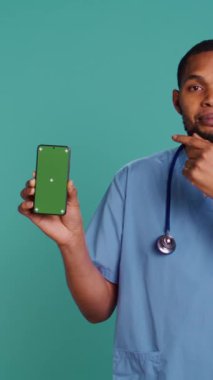 Afro-Amerikan hemşirelerin yeşil ekranlı cep telefonlarını gösteren dikey video görüntüleri. Gülümseyen sağlık uzmanı cep telefonunu, stüdyo geçmişini, kamera B 'yi tutuyor.