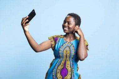 Stüdyoda akıllı telefon kamerasına gülümseyen Afrikalı Amerikalı kadın fotoğraf çekiyor ve sosyal medya uygulamalarına yüklüyor. Kadın model kendini mavi arka planda, etnik tarzda fotoğraflıyor..