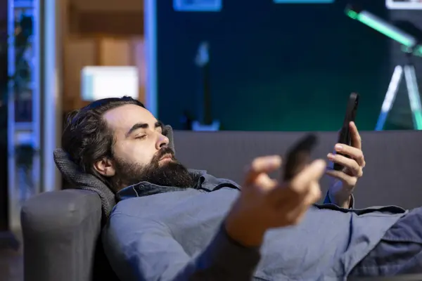 stock image Homeowner bored at home laying down, using TV to watch movies on streaming services and chatting with friends. Apathetic man lounged on couch, mindlessly consuming entertainment and using mobile phone