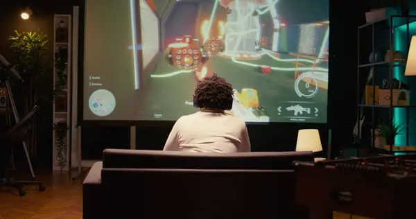 stock image Man watching gameplay footage of videogame competition on gigantic smart TV, relaxing on couch. BIPOC gamer in home theatre following gaming tournament on television set, zoom out shot