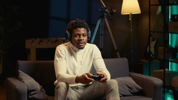 stock image Man in rgb lit apartment playing video games on couch, enjoying day off from work. Gamer battling enemies in online multiplayer shooter on gaming console, talking with teammates through headphones