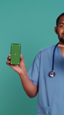 Neşeli erkek hemşirenin yeşil ekranlı akıllı telefonu işaret eden dikey video portresi. Renkli telefon tutan neşeli bir hastane çalışanı, mavi stüdyo arka planında izole edilmiş, kamera B