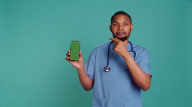 Afro-Amerikan erkek hemşirenin yeşil ekran cep telefonunu işaret eden portresi. Gülümseyen sağlık uzmanı cep telefonunu, stüdyo geçmişini, kamera B 'yi tutuyor.