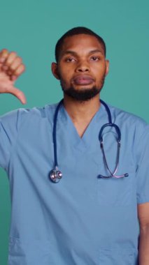 Dikey video üzgün Afrikalı Amerikalı erkek hemşire iş yerindeyken işaret veriyor, stüdyo arka planında izole bir şekilde. Mutsuz sağlık çalışanı önlük giyiyor onaylanmamış jestler yapıyor, kamera B