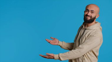Kameraya bir şeyin reklamını yapan gülümseyen kişi stüdyonun sol ya da sağ tarafını işaret ediyor. Orta Doğulu adam pazarlama kampanyası için reklam veriyor, mavi arkaplan pozu. Kamera 1.