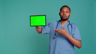 Neşeli hemşire krom anahtar tabletinde tıbbi talimat videosu gösteriyor. Hastane çalışanı yeşil ekran cihazında açıklayıcı sağlık bandı sunuyor, stüdyo arka planı, kamera B