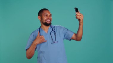 Erkek hemşire hastanede mesai arasında selfie çekmek için cep telefonu kullanıyor. Sağlık çalışanı fotoğraf çekmek için akıllı telefonu kullanıyor, stüdyo arka planında izole edilmiş, kamera B