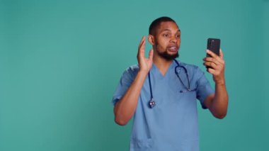Video konferansında cep telefonu kullanan, insanları selamlayan erkek hemşire. Stüdyo arka planında izole edilmiş, akıllı telefon kullanan hastalarla çevrimiçi teletıp görüşmesi yapan sağlık uzmanı, kamera B