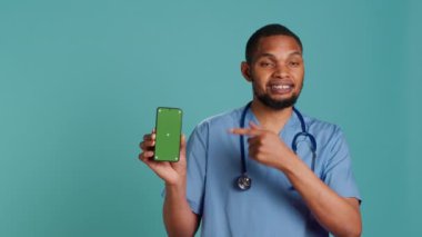 Afro-Amerikan erkek hemşirenin yeşil ekran cep telefonunu işaret eden portresi. Gülümseyen sağlık uzmanı cep telefonu, stüdyo arka planı, kamera A