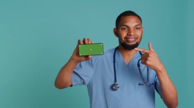 Neşeli erkek hemşirenin portresi izole edilmiş akıllı telefonu işaret ediyor. Radyasyon uzmanının elinde kroma anahtar telefon, mavi stüdyo arka planı, kamera A