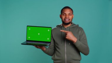 Yeşil ekran dizüstü bilgisayar tutan neşeli Afro-Amerikan adam tavsiye veriyor. Dost canlısı BIPOC çalışanı, model defteri işaret ederek, olumlu geri bildirim, stüdyo arka planı, kamera B