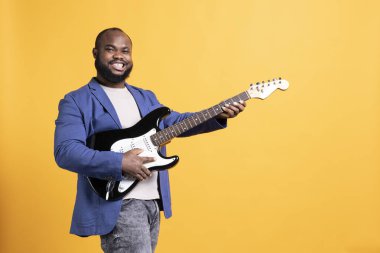 Gitar tutan mutlu adamın portresi, konser sırasında rock şarkıları söyleyen sarı stüdyo arka planında izole edilmiş. Neşeli sanatçı elektrikli müzik enstrümanı üzerinde beste yapıyor.