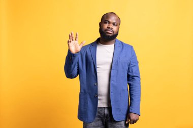 Afrikalı Amerikalı adam istemeyerek selamlama hareketi yapıyor. Duygusuz BIPOC çalışanının portresi. Birini selamlamak için kolunu kaldırıyor. Sarı stüdyo arka planında izole bir şekilde el kol hareketi yapıyor.