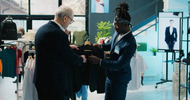 Afro-amerikan mağaza asistanı düzenli müşteriye alışveriş merkezinde bir takım elbise için yardım ediyor, kıdemli müşteri resmi kıyafetler almak istiyor. Yaşlı adam alışveriş merkezinde yeni koleksiyon için alışveriş yapıyor. Kamera B.