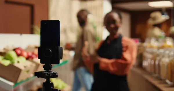 Stock image Market employee creating marketing commercial video to attract clients, promoting additives free pantry supplies in reusable jars and locally grown produce. Woman presenting bio products. Camera 2.