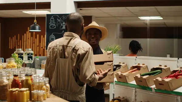 stock image Small business owner receiving new produce stock from contractor, bringing freshly harvested natural products from local farm. African american man supplying organic grocery store. Camera 1.