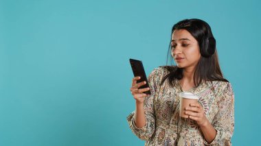 Joyous woman listening music, singing lyrics from phone screen, enjoying beverage. Cheerful person wearing headphones, listening songs, head bopping and holding coffee cup, studio background, camera A clipart