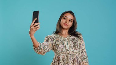 Neşeli Hintli kadın akıllı telefon kullanarak selfie çekiyor ve sosyal medyada yayınlıyor. Mutlu insan fotoğraf çekiyor. Fotoğraf makinesi kullanıyor. Stüdyo arka planında izole edilmiş.