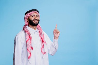 Geleneksel Arap kıyafetleri giyen gülümseyen Müslüman adam parmak stüdyosunun portresini göstererek ürününü sergiliyor. Neşeli İslami kişi yukarı çıkıyor ve kameraya bakıyor.