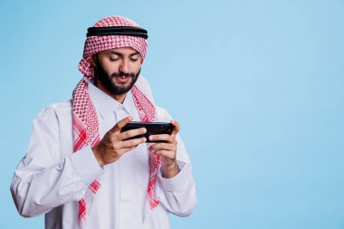 Geleneksel İslami kıyafetler giymiş gülümseyen Müslüman adam akıllı telefondan online oyun oynuyor. Arap kişi cep telefonu yazılım uygulaması ile stüdyoda sanal eğlencenin tadını çıkarıyor