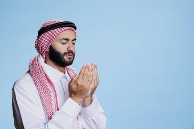 Müslüman kıyafetleri içinde kolları açık ve gözleri kapalı dua eden, ibadet ve inanç gösteren bir adam. Geleneksel başörtüsü ve cübbe takan Arap namazı, dini jestlerle ayakta duruyor.