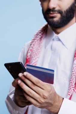 Geleneksel giysiler içinde, banka kartı ve cep telefonu kullanarak elektronik ödeme yapan Müslüman bir adam. Arap kişi internet mağazasından mal alıyor ve internetten ödeme yapıyor.