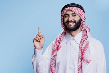 Arap kıyafetleri giymiş gülümseyen Müslüman adam yukarı doğru parmağıyla işaret ediyor ve kameraya bakıyor. Geleneksel bornoz ve başlık giymiş neşeli İslamcı kişi ortaya çıkıyor.