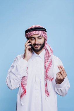 Müslüman elbiseli bir adam cep telefonuyla konuşurken koluyla gülümsüyor ve el kol hareketi yapıyor. Akıllı telefonla konuşurken ve stüdyoda dikilirken tobe ve ghutra giyen bir Arap.
