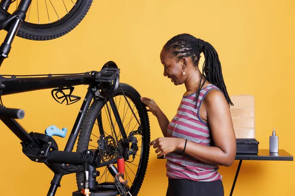 stock image Active young woman does yearly maintenance and adjustment of bicycle tire in front of isolated yellow background. Athletic african american lady examining her bike wheel for recreational cycling.