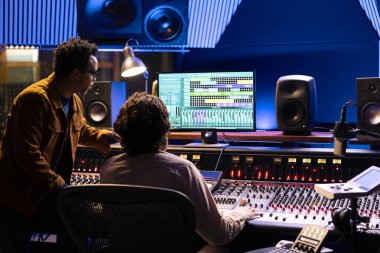 Şarkıcı ve yapımcı stüdyo kayıt istasyonunda ses panosu denetleyicisi üzerinde çalışıyor. Ses mühendisi müzik üretimi için dijital konsol kullanır, kontrol masasında faders ve anahtar düğmeleri üzerinde çalışır.