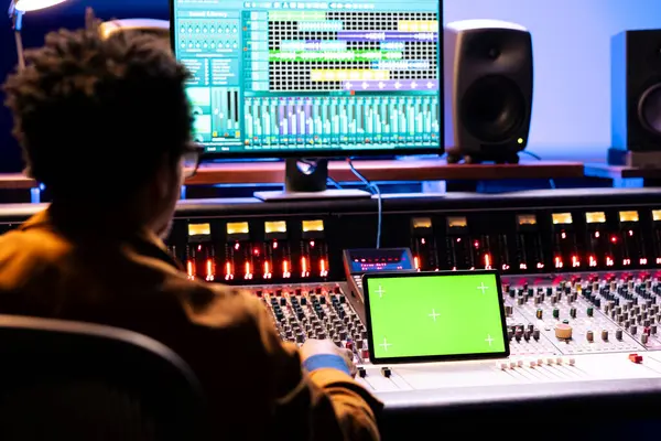 stock image African american audio engineer looks at greenscreen on device and digital editing software on computer, processing music in studio. Sound producer creating tracks and adjusting volume levels.