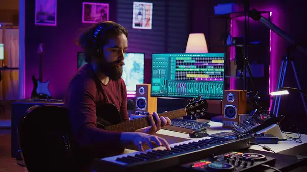 stock image Musical composer recording his guitar tunes and adding piano key sounds, creating a beautiful song with mixing console and daw software. Sound engineer working with electronic tools. Camera B.