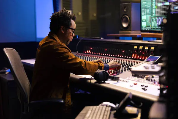 stock image African american tracking engineer editing tunes in control room, working with mixing console in professional recording studio. Young audio expert pushing faders and technical buttons.