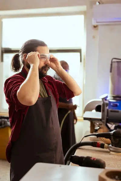 stock image Carpenter preparing to start production in furniture assembly shop, putting on safety glasses. Cabinetmaker at workbench ready to cut wood pieces, wearing protective equipment to avoid injuries