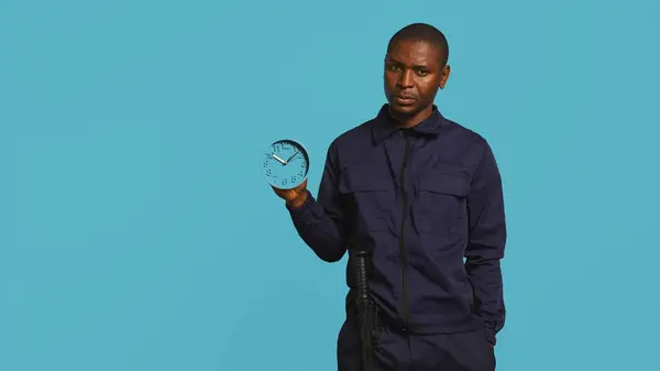 Stock image Security guard holding clock, rushing people out of property. Portrait of stern bodyguard showing visitors time is passing, preparing for closing down location, studio background, camera B