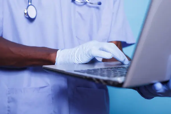 stock image Close-up of african american hands with gloves carrying and using digital laptop. Black medical nurse wearing blue scrubs and stethoscope typing on wireless computer.