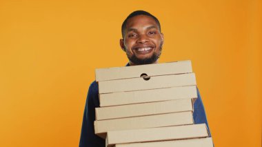 Kendine güvenen erkek kurye müşterilere fast food siparişi vermek için büyük pizza kutuları taşıyor. Stüdyoda paket servis ediliyor. Genç gülen teslimatçı dışarıdan yemek getiriyor. Kamera A.