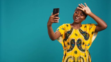 Özçekim için özgüvenle poz veren çekici doğal bir kadın mavi arka planda akıllı telefonuyla fotoğraf çekiyor. Genç adam stüdyoda fotoğraf çekiyor. Kamera B.