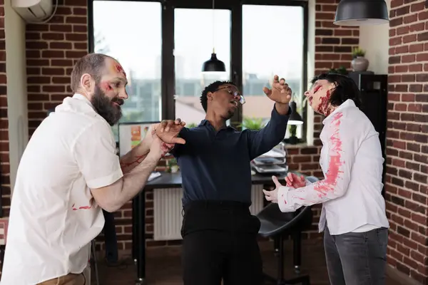 Stock image Employees wearing zombie costumes messing around with BIPOC manager in office. Team leader and workers dressed as reanimated corpses having fun during corporate Halloween party