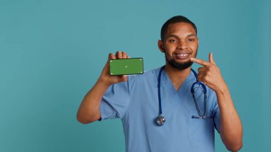 Neşeli erkek hemşirenin portresi izole edilmiş akıllı telefonu işaret ediyor. Radyasyon uzmanının elinde kroma anahtar telefon, mavi stüdyo arka planı, kamera A