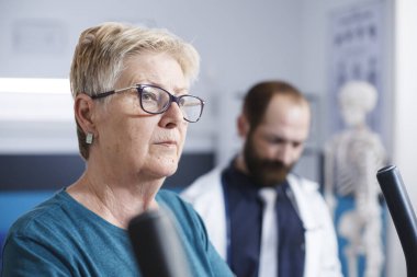 Kafkasyalı yaşlı bir kadının fizyoterapi için durağan bisiklet kullanırken çekilmiş bir portresi. Tıp doktorunun yardımıyla. Emekli hasta rehabilitasyon için hastanede..