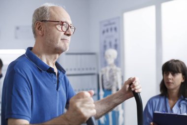 Mavi önlüklü kadın hemşirenin yardımıyla fizyoterapi için durağan bisiklet kullanan beyaz yaşlı bir adamın yan görüş portresi. Rehabilitasyon için hastanedeki kıdemli hasta.