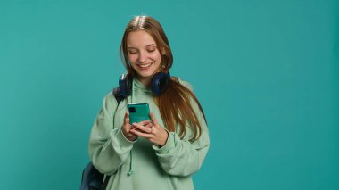 Portrait of happy teenager reading messages on phone, delighted after receiving good news. Cheerful woman excited by SMS on smartphone, celebrating, studio background, camera A clipart