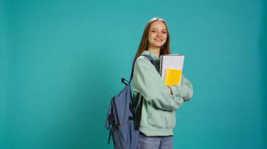 Sırt çantasında okul defteri ve notları olan gülümseyen kadın portresi stüdyo arka planında izole edilmiş. Okul malzemeleriyle neşeli bir kız, üniversiteye gitmeye hazırlanıyor, kamera B
