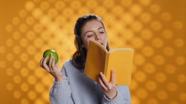 Kitap okuyan ve taze elmanın tadını çıkaran, sağlıklı yaşayan, stüdyo arka planında izole edilmiş bir kadın. Yeşil meyve yiyen, biyo-atıştırmalıkların tadını çıkaran kitap kurdu, kamera B