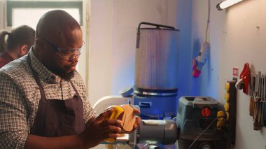 Stüdyoda BIPOC çalışanı, arka planda ahşap yüzeyi zımparalamak için zımpara kağıdı kullanıyor. Tahta işçiliği uzmanı odun parçasının, kamera B 'nin zararlarını gidermek için aşındırıcı sünger kullanıyor.