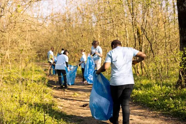 stock image African american woman grabbing junk and plastic waste from the forest, doing voluntary work to preserve and protect the environment. Female activist using claw tool to collect garbage and junk.