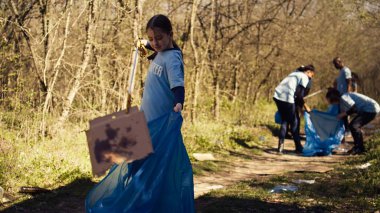Küçük çocuk ormanda çöpleri toplamak ve toplamak için maşa kullanıyor, doğal bir habitattan çöp topluyor. Çevresel bakım ve plastik atıklarla büyüyen küçük bir kız. Kamera B.