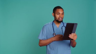 Video konferansında erkek hemşire tablet kullanıyor, insanları selamlıyor. Hastalarla çevrimiçi teletıp görüşmesi yapan sağlık uzmanı. Mavi stüdyo arka planında izole edilmiş. Kamera B.