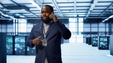 Bilgi işlem teknisyeni veri merkezinde telefonla konuşuyor, sunucu bakımını ve sorun çıkarma görevlerini koordine ediyor. Sunucu rafları ve yüksek teknoloji ekipmanları ile çevrili bir yönetici. Telefon görüşmesi, kamera A